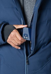 Marineblaue Jacke mit einem Reißverschluss und einem grauen Kragen. Eine Hand zieht ein blaues Smartphone aus der Innentasche, das mit einer Dual-Kamera ausgestattet ist.