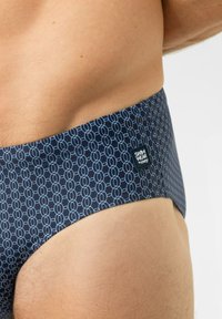 Slips de bain bleu marine avec un motif géométrique bleu clair, dotés d'une étiquette carrée sur la ceinture. Texture lisse, coupe ajustée.