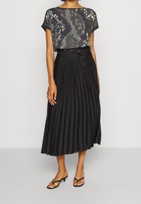 Černá skládací sukně s vysokým pasem a bočními knoflíkovými detaily, spárovaná se šedou vzorovanou blůzkou s žlutými a modrými akcenty.