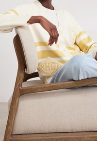 Cardigan crema con strisce gialle, tasche frontali e logo ricamato. Texture morbida. Modella seduta su una sedia texturizzata beige chiaro.