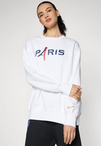 Biela mikina s nápisom "PARÍŽ" v modrej a červenej farbe, so zlatým logom Nike na rukáve. Má voľný strih a ribovanými manžetami.