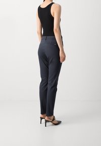 Kobieta ubrana w ciemnogranatowe, dopasowane spodnie, czarny top bez rękawów i czarne mules na wysokim obcasie, z widocznym tatuażem na kostce, stoi na białym tle.