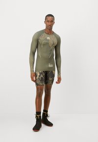 Olivegrünes Langarm-Kompressionsshirt mit Blättermuster und weißen Akzenten, kombiniert mit gemusterten schwarzen Shorts und schwarzen Sportstiefeln.