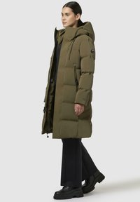 Parka lunga color verde oliva, imbottita e con cappuccio, dotata di tasche con zip e una silhouette leggermente affusolata. Realizzata in materiale morbido e resistente alle intemperie.
