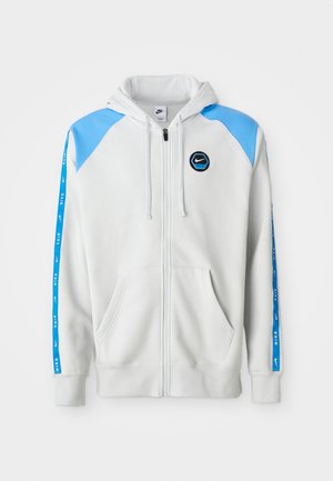 Svetlo siva zip-up dukserica z modrimi naglasnimi detajli in logotipom, ki ima dva sprednja žepa in modre črtaste detajle vzdolž rokavov.