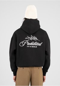 Sudadera negra de gran tamaño con texto bordado en blanco "Prohibido 10119 BERLÍN" y un diseño de hoja en la parte trasera. Puños acanalados y material de felpa visible.