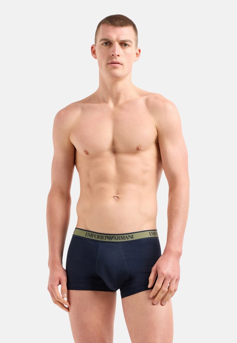Boxer shorts de algodón azul marino con una textura suave, que presentan una cinturilla verde en contraste etiquetada como "Emporio Armani". Diseño ajustado, cobertura mínima.