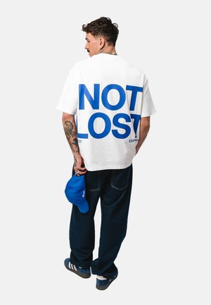 Mężczyzna z tatuażami nosi białą oversize koszulkę z dużym niebieskim napisem "NOT LOST" na plecach, trzyma niebieską czapkę, stoi na jednolitym tle.