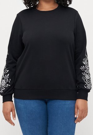 Czarna bluza z długim rękawem z białą kwiatową haftem na rękawach, noszona przez osobę o kręconych włosach i niebieskich dżinsach, stojącą na białym tle.