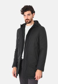Cappotto lungo nero con cappuccio, dotato di chiusura frontale a zip e automatici, presenta tasche laterali e una texture liscia e opaca. Il modello posa su uno sfondo neutro.