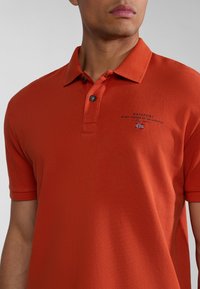 Oranžová polo košile z texturované látky, s límečkem, dvěma knoflíky a malým vyšitým logem s vlajkou na hrudi.