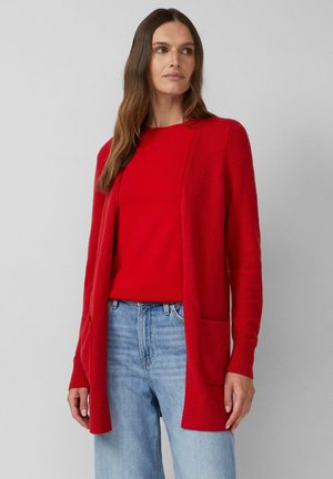 Frau mit langen braunen Haaren, die eine rote Strickjacke und ein passendes rotes Oberteil zu hellblauen Jeans trägt, steht vor einem schlichten grauen Hintergrund.