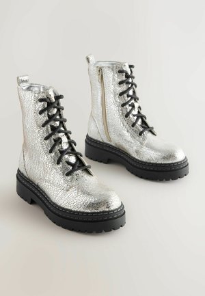 Ett par silverfärgade metallic stridsstövlar med spruckna texturer, svarta tjocka sulor, svarta snören och sidizippar på en ljus bakgrund.