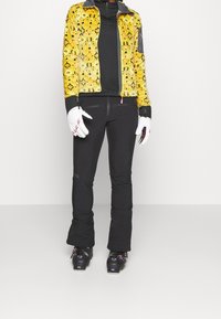 Chaqueta amarilla con patrón y detalles grises, camiseta de manga larga negra, pantalones de esquí negros, guantes blancos y botas de esquí negras con detalles rosas.