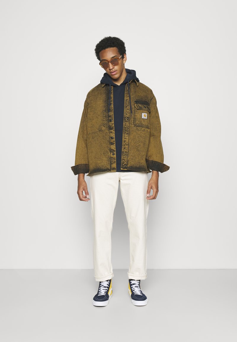 Braune Denimjacke mit einer Tasche, schwarzes Kapuzensweatshirt, cremefarbene Hose und marineblaue High-Top-Sneaker mit gelben Akzenten. Braune Sonnenbrille.
