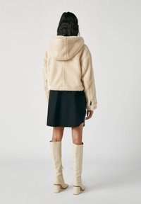 Kobieta z ciemnymi falującymi włosami, ubrana w beżowy, krótszy polarowy hoodie, czarną mini sukienkę i kremowe kozaki do kolan, stojąca na tle jednolitym.