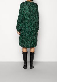 Vestido verde con estampado, de mangas largas, escote redondo y corte suelto. Combinado con botas negras brillantes hasta la rodilla.