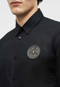 Άνδρας που φοράει μαύρο πουκάμισο με κουμπιά, με στρογγυλό έμβλημα Versace Jeans Couture στο αριστερό στήθος.