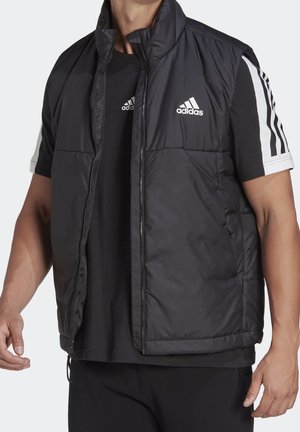Homme portant une veste matelassée sans manches noire Adidas par-dessus un t-shirt noir Adidas avec des manches blanches à trois bandes, debout sur fond blanc.
