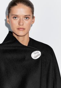 Žena s vlasy stáhnutými dozadu nosí černé oblečení připnuté velkou hladkou oválnou stříbrnou broží na bílém pozadí.