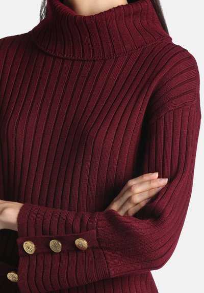 Femme portant un pull à col roulé côtelé bordeaux avec des boutons dorés sur le poignet, bras croisés sur la poitrine.