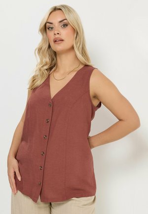 Lyshåret kvinde iført ærmeløs, knap-op, rustfarvet top og beige bukser, stående med den ene hånd på hoften mod hvid baggrund.