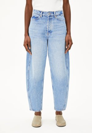 Personne portant un jean ample bleu clair taille haute, un haut blanc et des chaussures beiges sans lacets, debout devant un fond blanc uni.