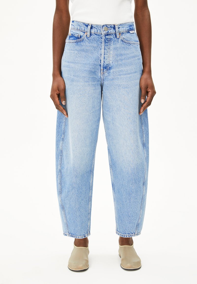 Personne portant un jean ample bleu clair taille haute, un haut blanc et des chaussures beiges sans lacets, debout devant un fond blanc uni.