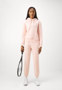 Felpa rosa chiaro e pantaloni jogger a sigaretta realizzati in morbido tessuto, con tasca frontale e polsini a coste, abbinati a una racchetta da tennis e sneakers.