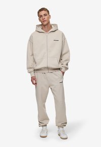 Beige hættejakke med lynlås og matchende sweatpants, lavet af blødt materiale. Har store forlommer og subtile logo-detaljer på brystet og låret.