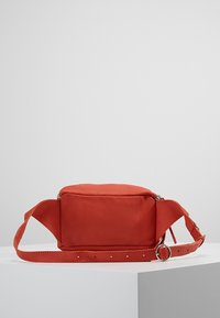 Bolso bandolera de ante rojo con forma rectangular, correa ajustable y herrajes plateados. Cierre de cremallera en la parte superior y textura suave.