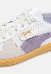 Gros plan d'une basket blanche Puma Palermo avec des accents en suède beige et violet, des lacets blancs et une semelle en caoutchouc naturel.