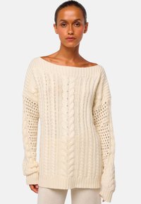 Cremfarbener Strickpullover mit weitem Halsausschnitt, strukturiertem Zopfmuster und offenknotigen Ärmeln. Weicher, dicker Stoff und gerippte Bündchen.