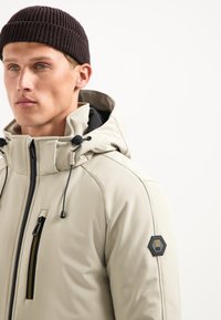 Lichtbeige gewatteerde jack met een gladde afwerking, zwarte ritssluiting zak, verstelbare trekkoorden en een hexagonaal logo-patch op de mouw.