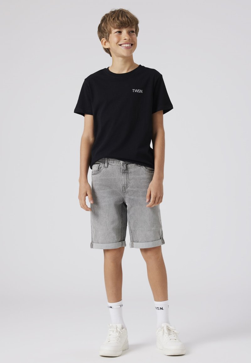 T-shirt en coton noir avec logo blanc, associé à un short en denim gris retroussé au bas, et des baskets blanches à semelles épaisses.