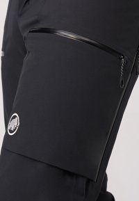 Zwarte geïsoleerde broek met een slank ontwerp, voorzien van een ritszak op de dij en een geborduurd logo in het wit. Gestructureerde stof met strategische stiksels.