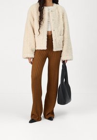 Krämmfärgad faux fur-jacka med en boxig passform, kombinerad med bruna ribbade vida byxor. Svarta spetsiga skor och en svart tote-väska.
