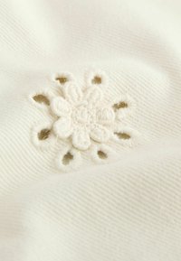 Tissu blanc avec un motif de fleurs brodées ; le centre présente une fleur texturée de couleur crème entourée de petits trous ouverts dans le tissu.