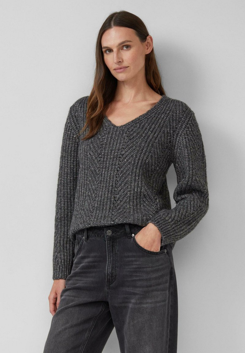 Grau gestrickter Pullover mit V-Ausschnitt, strukturiertem Muster und langen Ärmeln, kombiniert mit dunkelgrauen Jeans, die eine entspannte Passform zeigen.