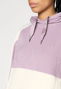 Μωβ φούτερ με λευκές λεπτομέρειες, που διαθέτει ένα μεγάλο κεντητό λογότυπο διαγωνίως στο στήθος, κουκούλα με κορδόνι και μαλακή, λεία υφή.