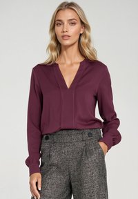 Bordowa bluzka z dekoltem w kształcie litery V i długimi rękawami, z delikatnymi detalami szycia. Połączona z chropowatymi, szaro-wysokim spodniami.