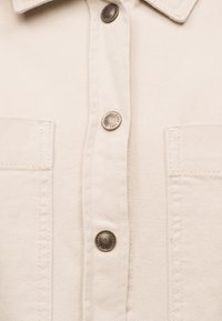 Κοντινή λήψη του εμπρός μέρους μπεζ τζιν μπουφάν με μεταλλικά κουμπώματα και δύο τετράγωνες τσέπες στο στήθος με λεπτομέρειες ραφής.