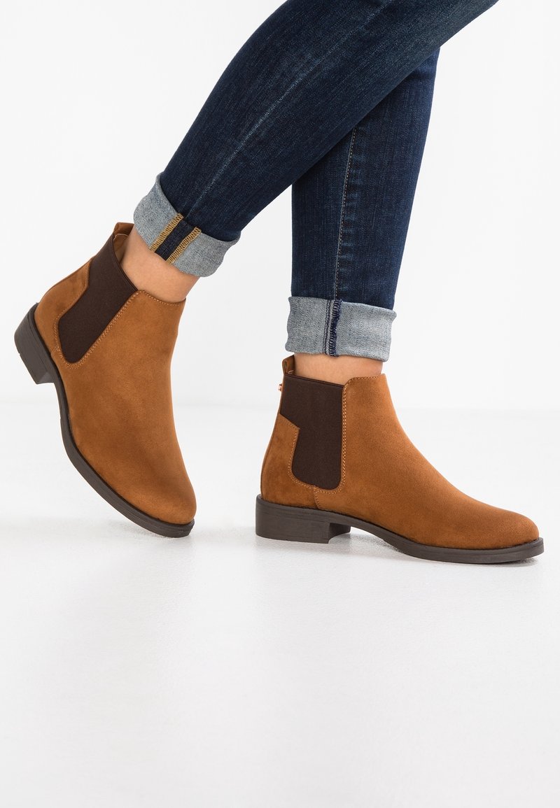 Botines de ante marrón con paneles elásticos en los lados y un tacón bajo, combinados con jeans oscuros de denim con dobladillo. Los botines tienen una textura suave.