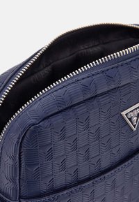 Sac à dos synthétique bleu marine texturé avec un motif en diamant, fermeture éclair argentée et détail de logo, avec un intérieur en tissu foncé.