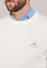 Felpa bianca con collo a girocotone a coste, dotata di un piccolo logo ricamato in blu e beige sopra il lato sinistro del petto.