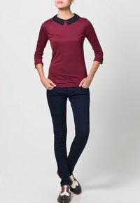 Top ajustado en color burdeos con cuello negro, de manga larga y detalles de botones en los puños, combinado con jeans ajustados azul oscuro y zapatos con cordones.