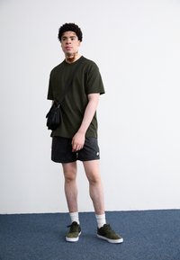 Olivenfarvet t-shirt, sorte mønstrede shorts, hvide crew-sokker og grønne ruskinds sneakers. Lille sort taske slidt over skulderen.
