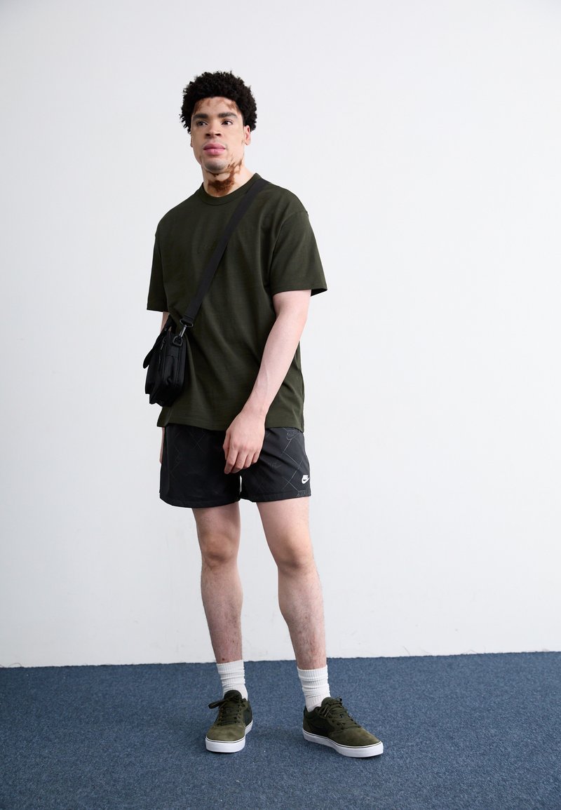 Olivgrünes T-Shirt, schwarze gemusterte Shorts, weiße Crew-Socken und grüne Wildledersneaker. Kleine schwarze Tasche über die Schulter gehängt.