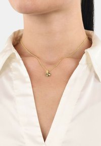 Collana d'oro con un piccolo pendente di pietra verde, posizionata su una camicetta bianca con scollatura a v. La catena ha un design delicato.