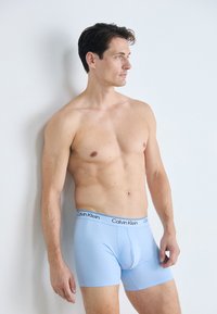 Man met kort donker haar in lichtblauwe Calvin Klein boxer shorts, die zonder shirt tegen een witte muur staat en naar rechts kijkt.